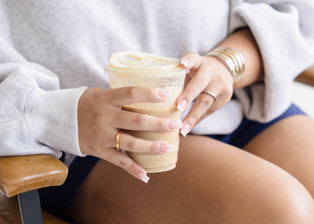 A woman sitting in a wooden rocking chair wearing a light gray off-the-shoulder sweatshirt and holding a takeaway coffee cup.