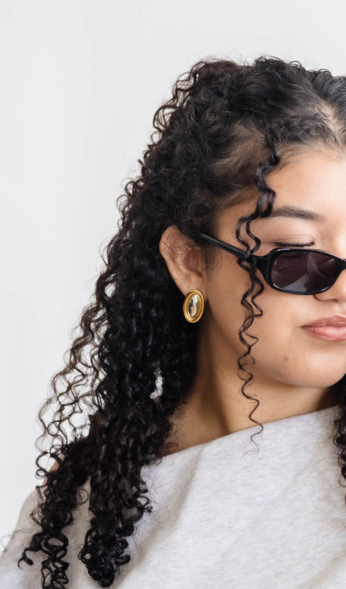 A woman with curly dark hair wearing black cat-eye sunglasses and a light gray oversized sweatshirt, posing against a plain white background.